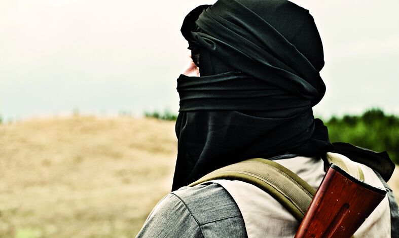 Eine Person mit einem schwarzen Kopftuch und einer Waffe auf dem Rücken steht in einer ländlichen Umgebung. Der Blick ist nach hinten gerichtet, während der Hintergrund eine hügelige Landschaft zeigt.